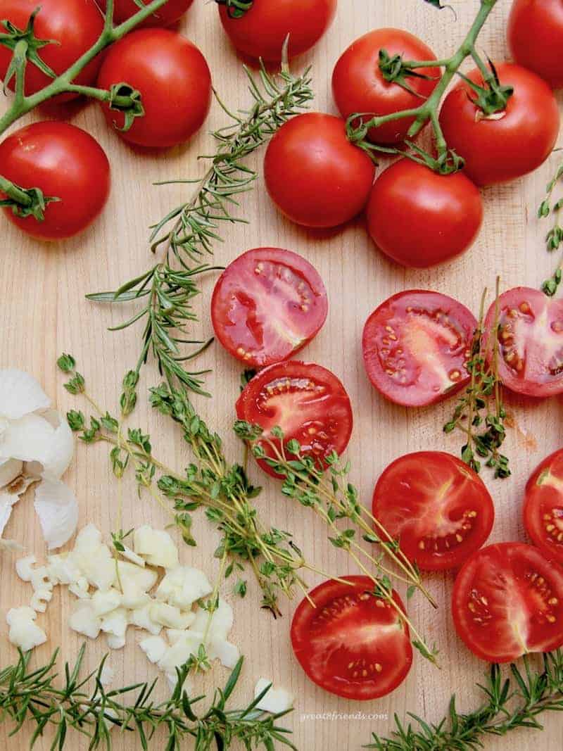 Fresh tomatoes, rosemary, oregano, and garlic on a wood cutting board.