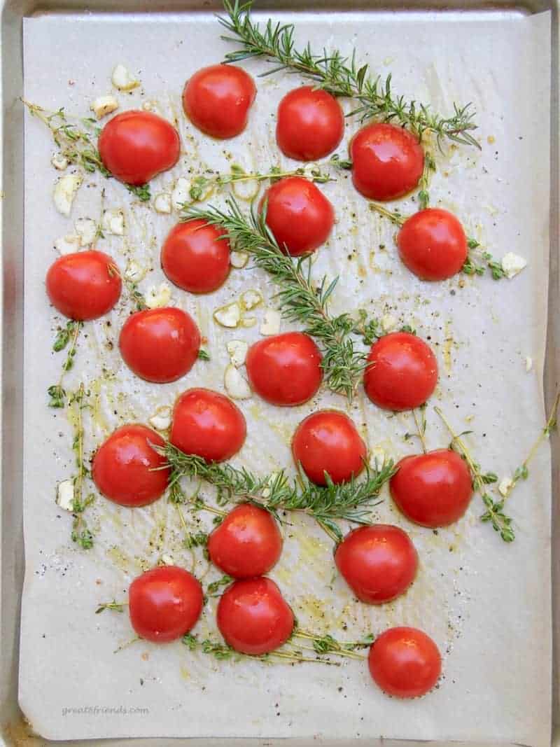 Fresh tomatoes, rosemary, thyme, and garlic on a parchment covered sheet pan ready for roasting.