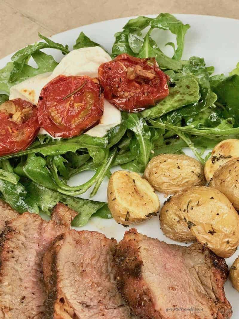 Upclose photo of roasted tomato and arugula salad on a dinner plate with roasted potatoes and slices of tri-tip roast.