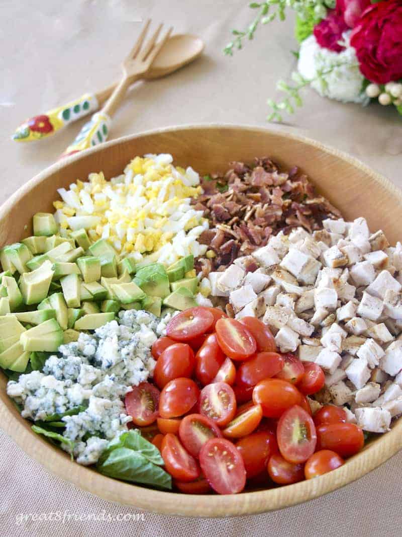 A cobb salad in a wooden bowl with salad servers in the background.