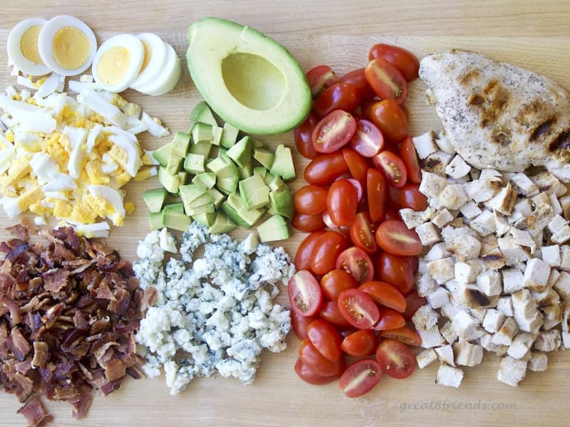 Cutting Board with chopped egg, bacon, avocado, bleu cheese, cherry tomatoes halved and cubed chicken.