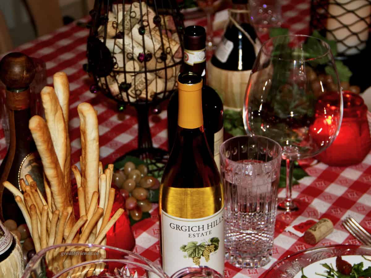 A table setting with wine, breadsticks and a red and white tablecloth for an Italian dinner party.