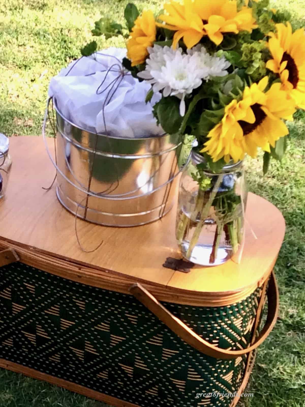 An old fashioned picnic basket with a tin buchet and vase of sunflowers on top.