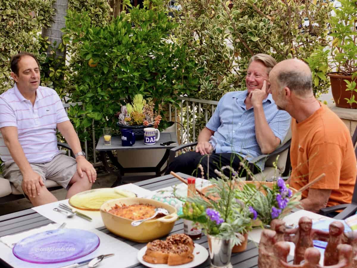 Three men sitting on a patio with a table of food in the foreground.