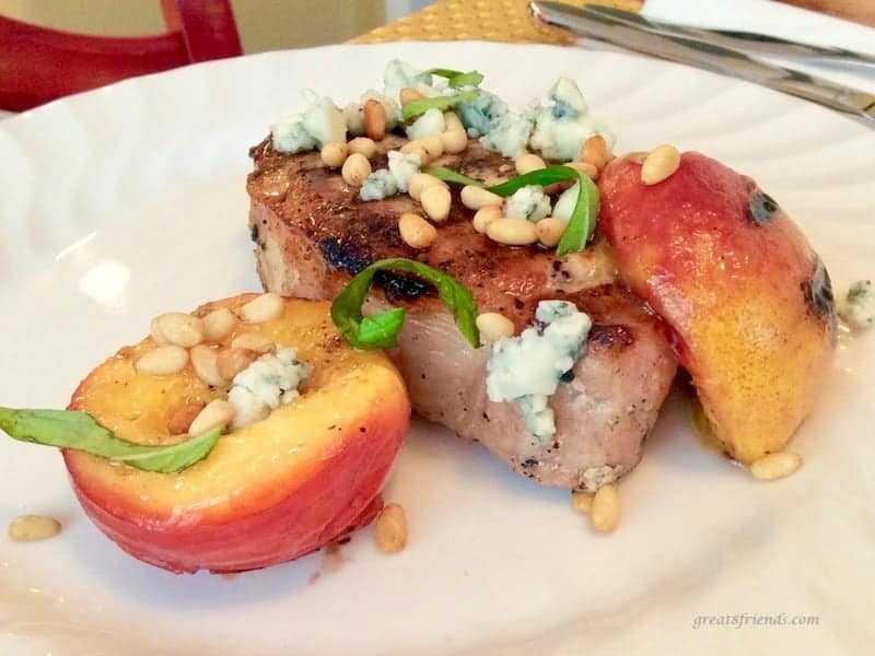 A grilled pork chop is served on a white plate with grilled nectarines, crumbled blue cheese and pine nuts.