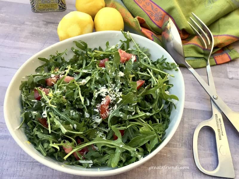 Arugula Salad with Lemon Parmesan Dressing in bowl with salad tongs, zested lemons and green and orange towel.