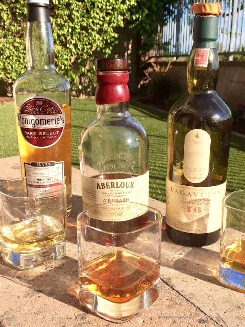 Three bottles of Scotch Whisky with a glass in front of each sitting on a stone in a yard.