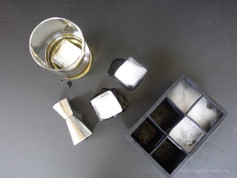 Overhead shot of large ice cubes, some in black silicone cube tray and some outside. with a glass and a shot glass.