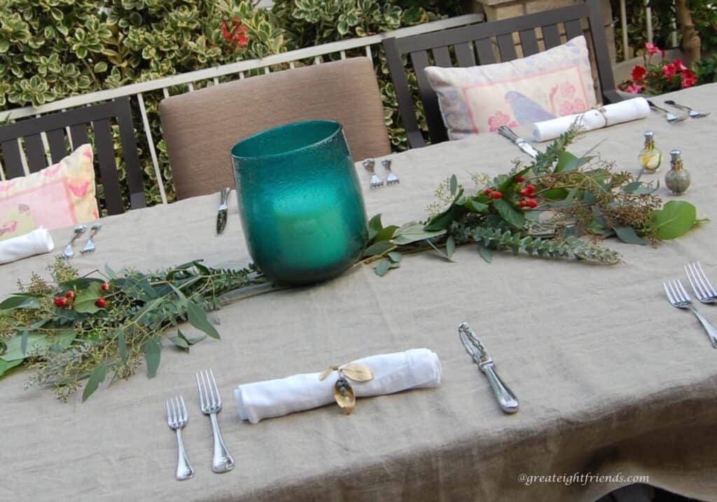 A table set with a blue glass hurricane in the center and greenery.
