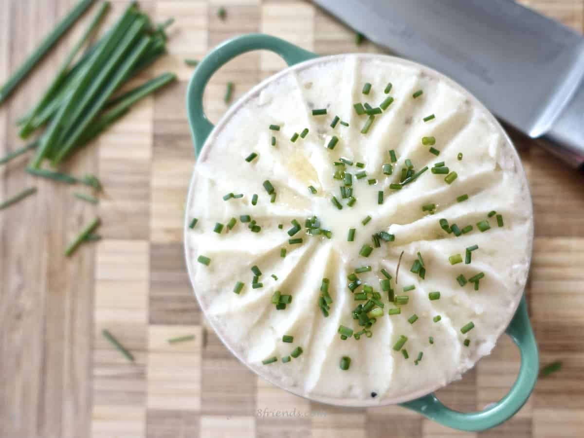 Overhead shot of Pommes de Terre Robuchon (mashed potatoes) in a dish with green handles topped with chopped chives. It's sitting on a checkered cutting board with a butcher knife to the right and some unchopped chives to the left.