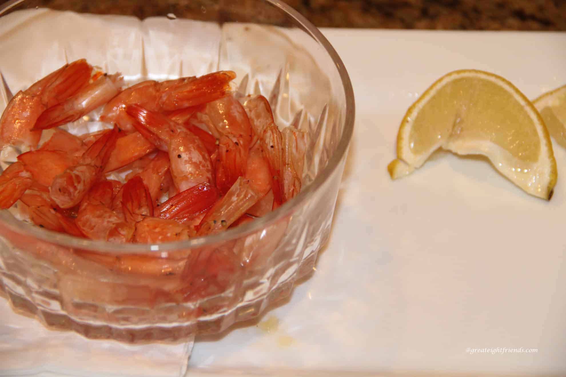 Shrimp tails in bowl on an empty plate with two lemon wedges on the side.