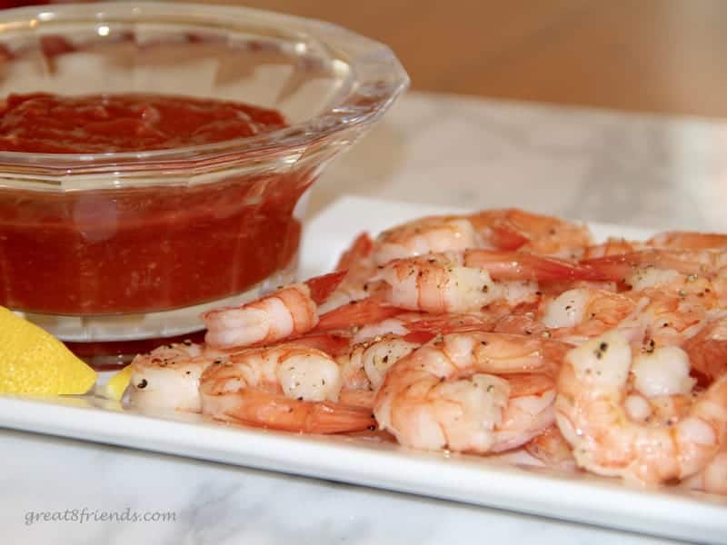 Shrimp and cocktail sauce on a white platter with a wedge of lemon on the side.