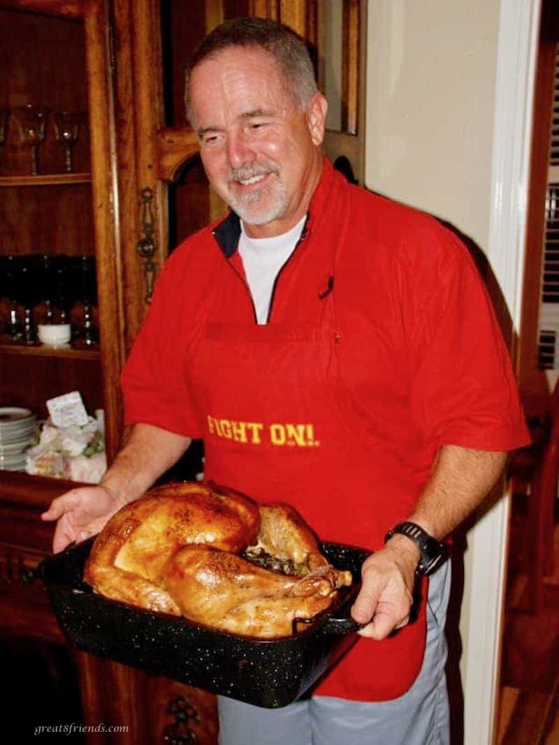 A man with a red apron on holding a roasting pan with a roasted turkey in it.