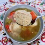 Upclose photo of a matzo ball in a spoon with the soup in a bowl below.