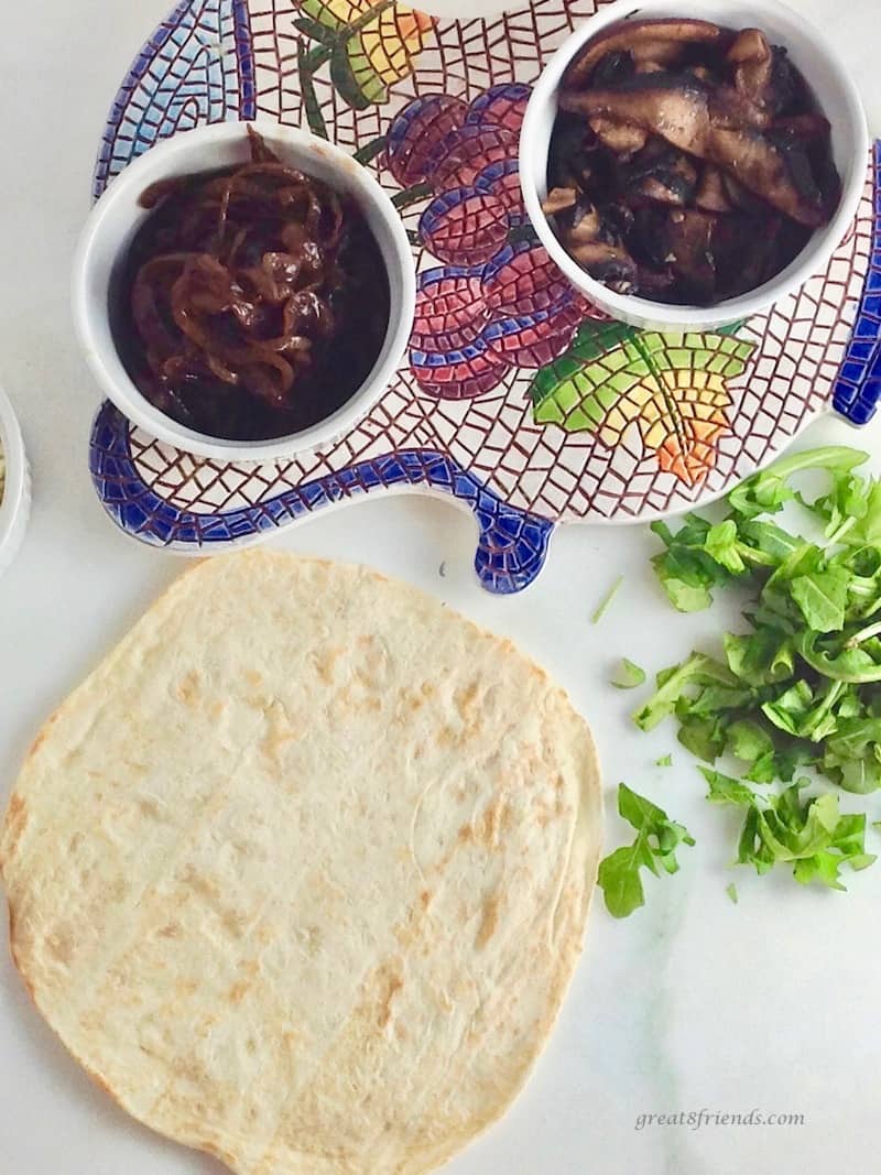 Overhead shot of flour tortillas, caramelized onions in small bowls, chopped arugula and sauteed mushrooms.