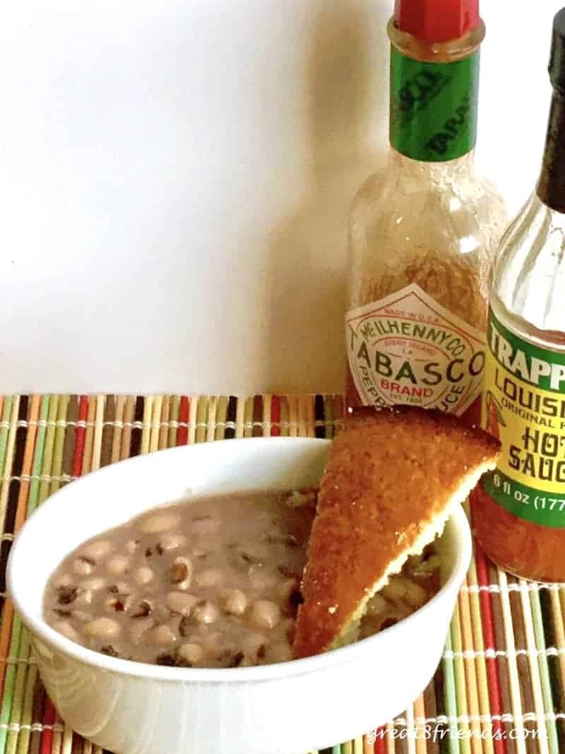 A white bowl of black eyed peas with a wedge of cornbread sitting on the side and 2 bottles of hot sauce in the background.