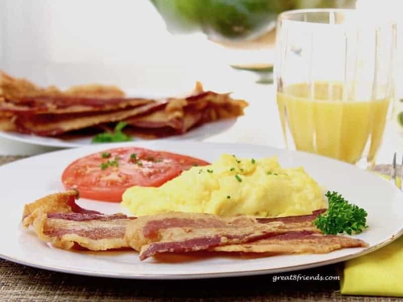 Slices of bacon, scrambled eggs and a slice of tomato on a plate with a glass of orange juice and more slices of bacon in the background.