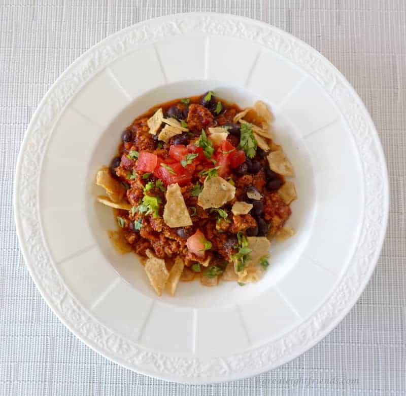 Overhead shot of chili in a white bowl topped with fresh tomatoes, cilantro, and broken tortilla chips.