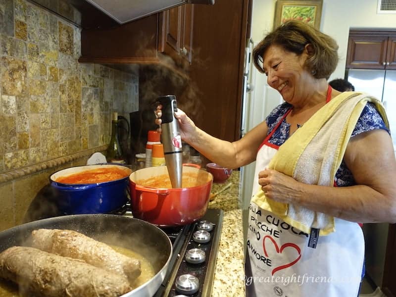 An Italian woman cooking red sauce and beef braciole at the stove using an immersion blender in one red pot.