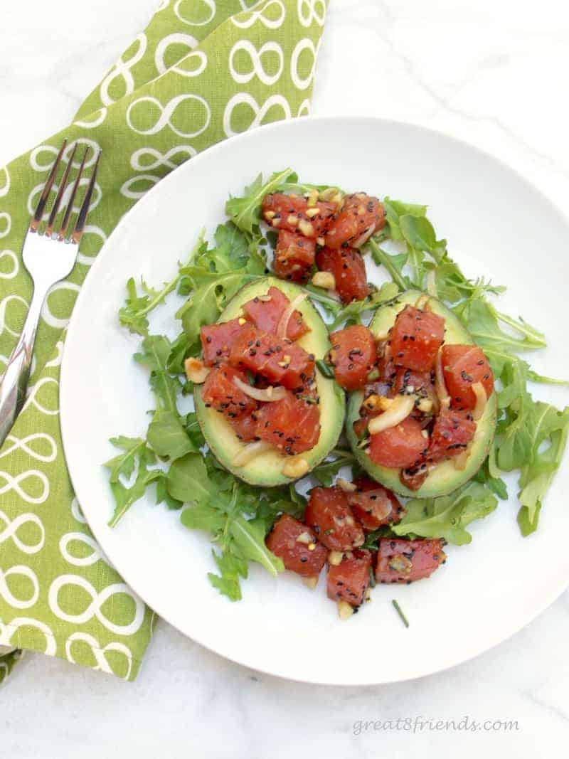 A white plate with avocado halves facing up with fresh ahi poke in each of two halves with arugula on the plate as well. A fork and a napkin are on the side.