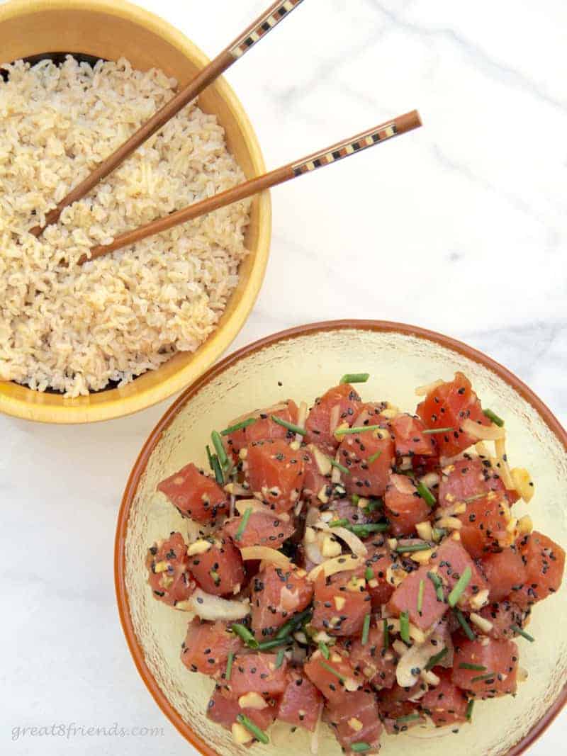 Fresh ahi poke salad in a glass bowl with another wooden bowl along side with brown rice and chop sticks.