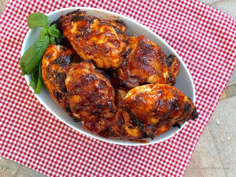 Photo of pieces of barbecued chicken in a white oval dish sitting on a red and white checked tablecloth. The dish is garnished with a sprig of basil leaves.