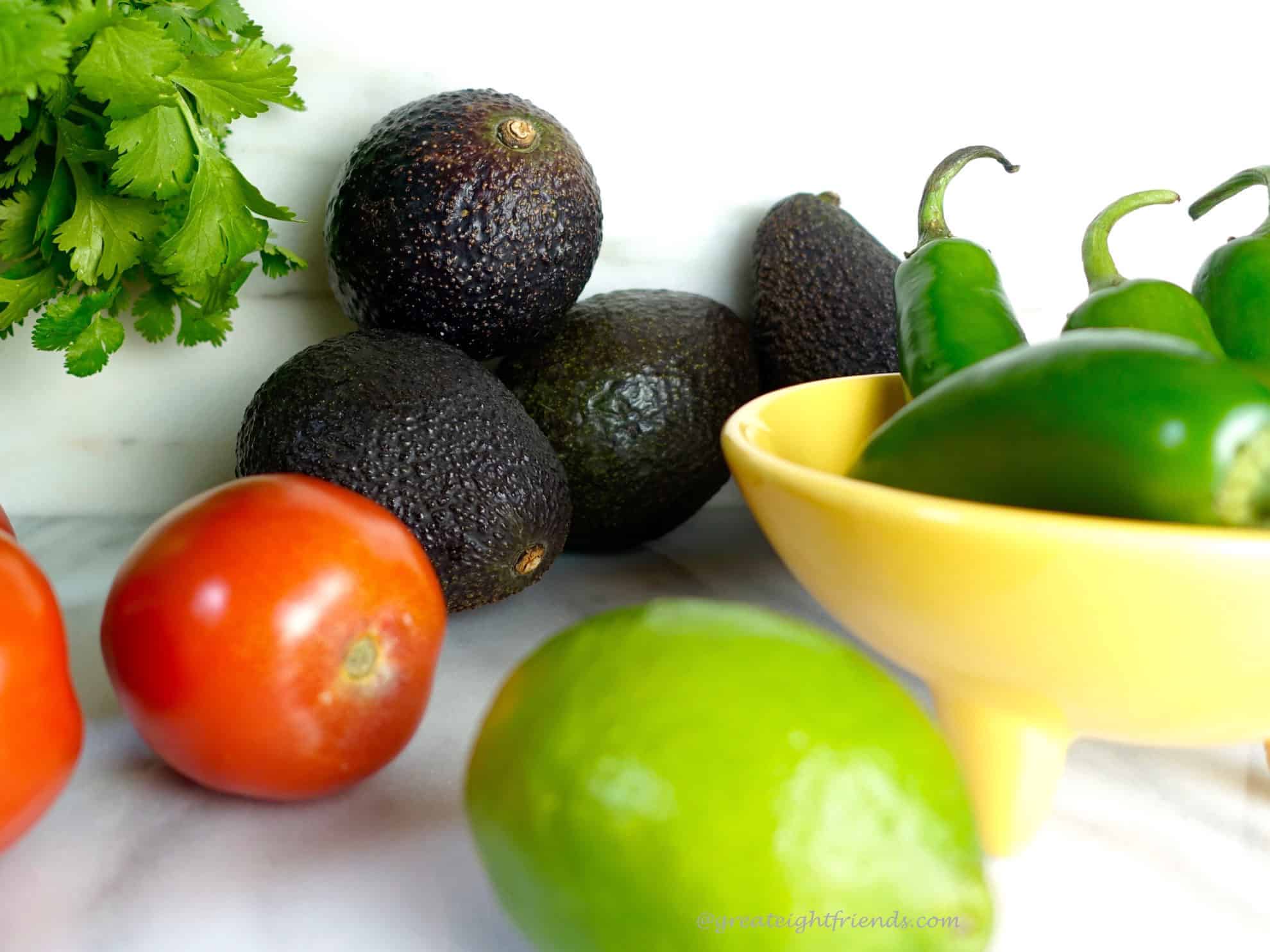 Avocados, cilantro, jalapenos in a yellow bowl, tomatoes and a lime.