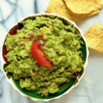 Overhead shot of a bowl of guacamole topped with a wedge of tomato with tortilla chips scattered around.