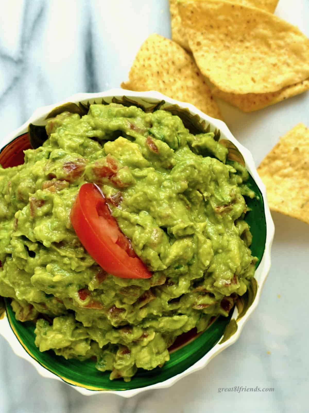 Overhead shot of a bowl of guacamole topped with a wedge of tomato with tortilla chips scattered around.