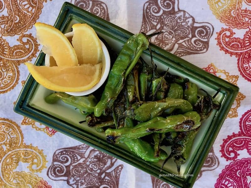 Overhead shot of a rectangular plate holding charred shishito peppers with some lemon wedges.