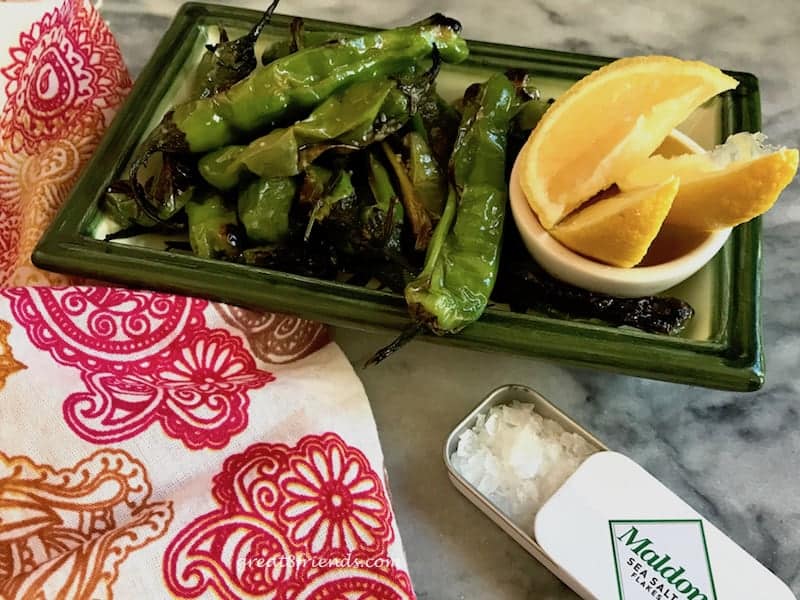 A green rectangular plate with charred shishito peppers, lemon wedges and a small box of flaky salt.