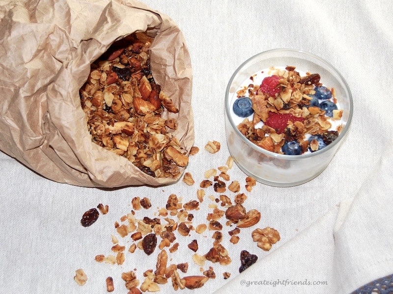 A brown paper sack with granola spilling out of it and a small glass bowl with yogurt and granola and berries in it.