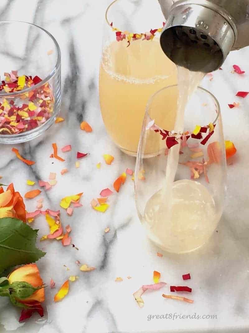 Pouring cocktail into stemless flutes rimmed with flower confetti.