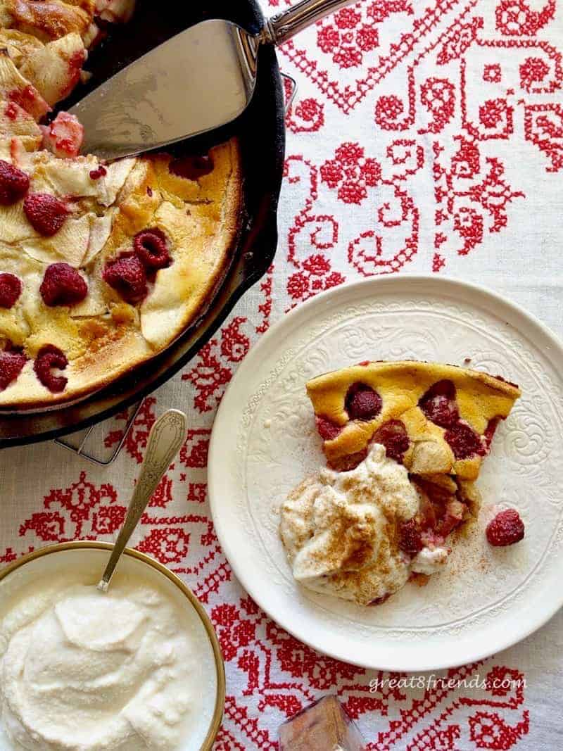 Overhead shot of a slice of Apple Berry Dutch Baby on a white plate next to the pan of Dutch Baby with a bowl of ricotta cream.