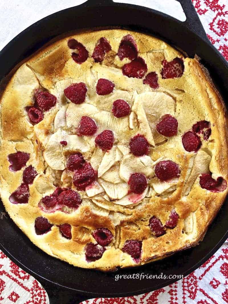 Overhead vertical shot of Apple Berry Dutch Baby in cast iron pan on a white tablecloth with red embroidery.