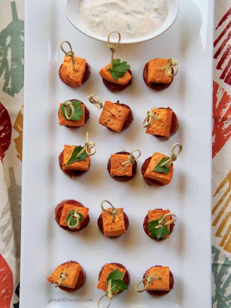 Several appetizer sticks on a white rectangular platter with a sweet potato piece and slice of sausage on the end.