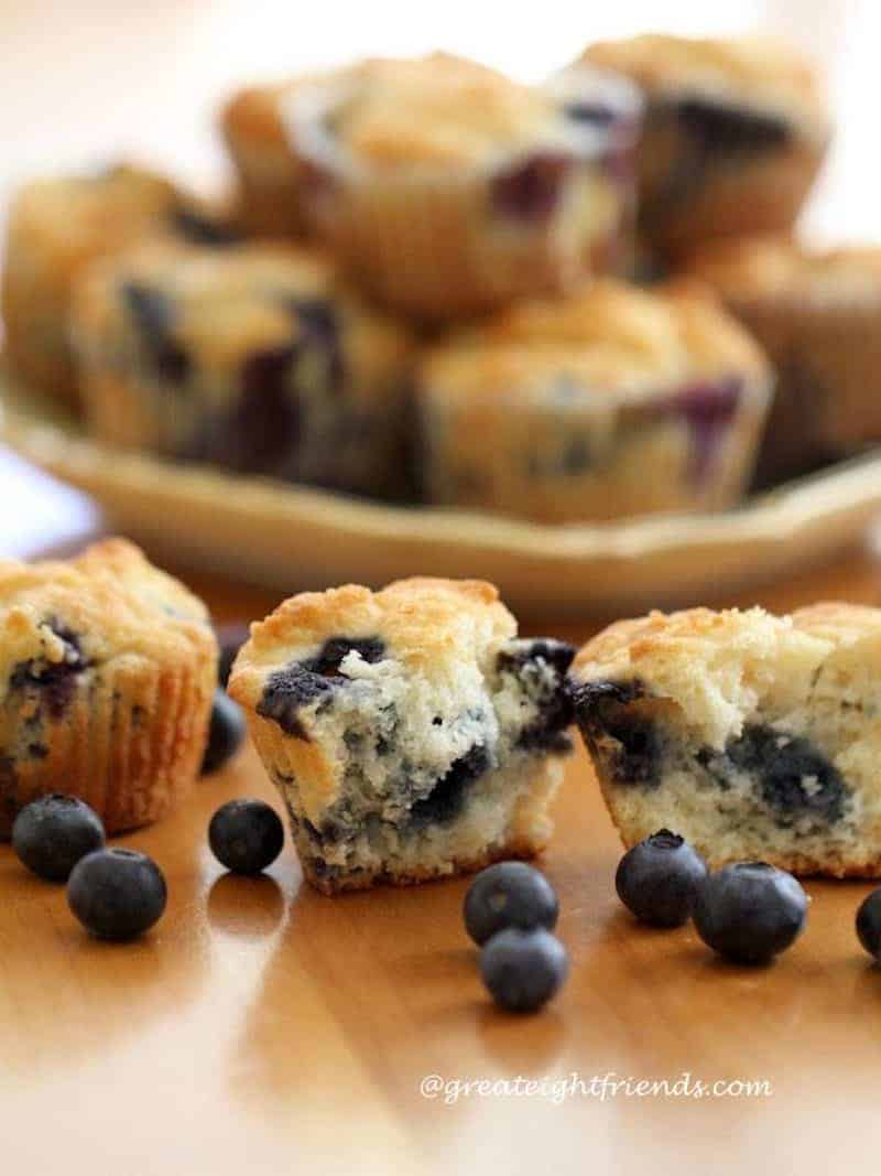 Homemade blueberry muffins on a plate in the background with a few on the table in the front broken in half with a few fresh blueberries on the table.