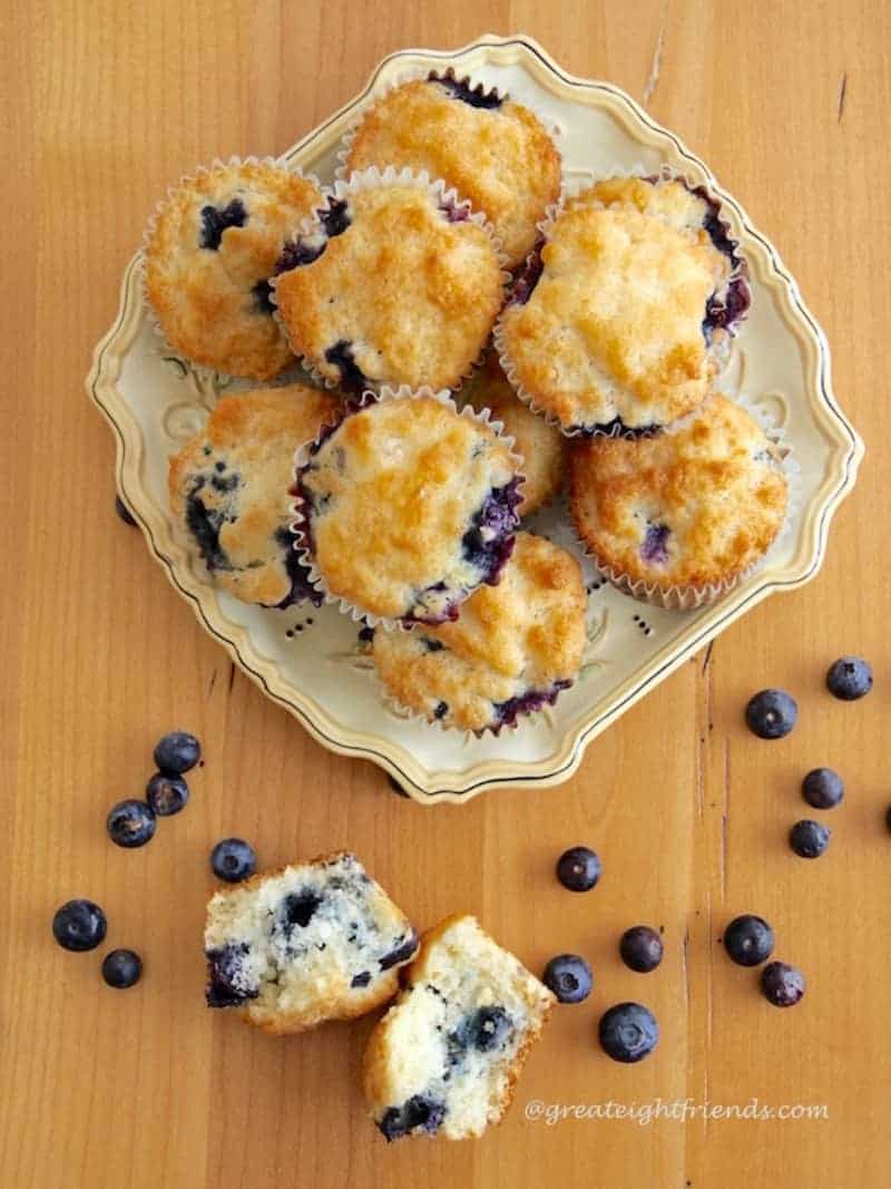 Over head shot of Homemade blueberry muffins on a plate with one on the table that has been broken into two pieces with fresh blueberries on the table.