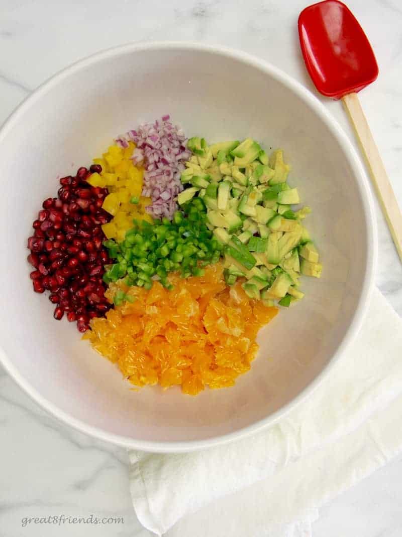 All the ingredients needed to make a Christmas salsa; pomegranate seeds, chopped oranges, cut avocado, cut green pepper, cut red onion in a large white bowl.
