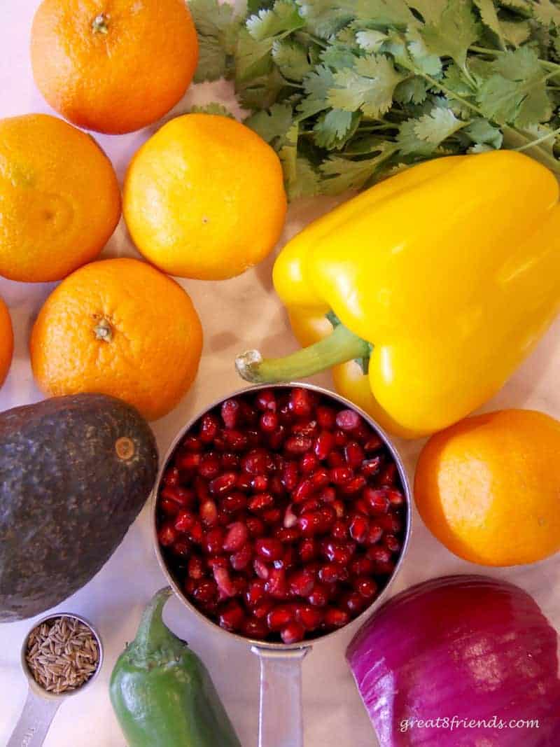 Overhead shot of all the ingredients needed to make a salsa including pomegranate seeds, whole avocado, clementines, red onion, jalapeño, cilantro and cumin seeds.