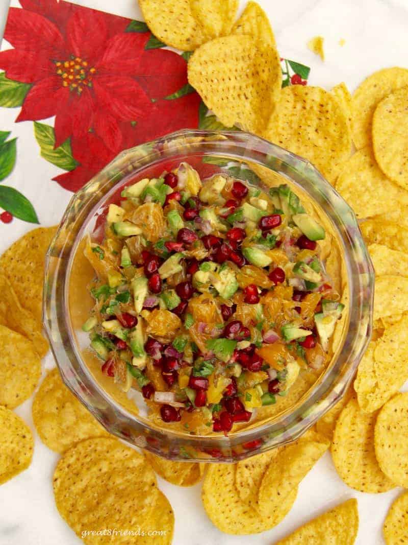 Overhead shot of a bowl of avocado salsa with pomegranate seeds with tortilla chips all around.