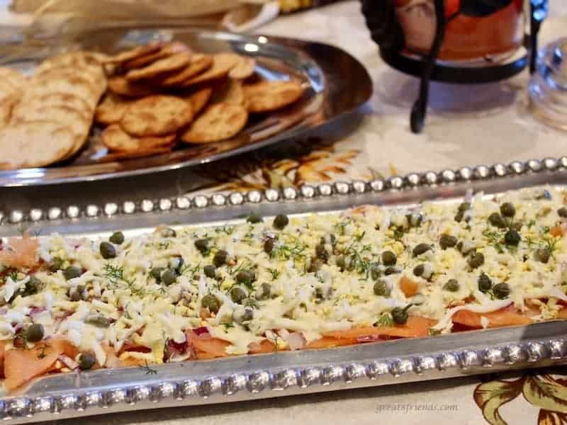 A decorative rectangular silver tray with smoked salmon layered dip with crackers in the background.