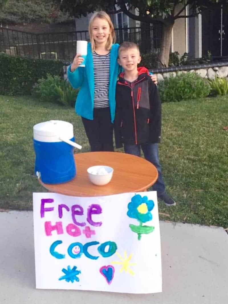 Two children outside standing behind a round wood table with a jug on top and a sign saying "Free Hot Coco."