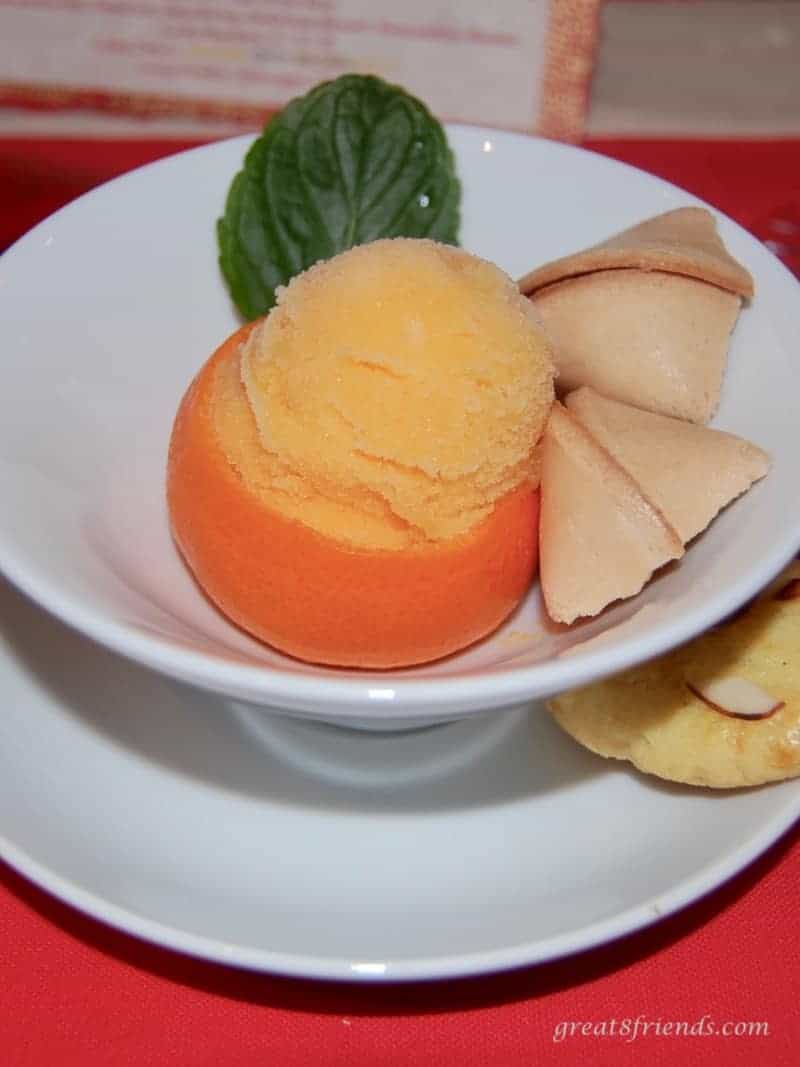 A scoop of tangerine sorbet in a tangerine shell with a fortune cookie and a leaf.