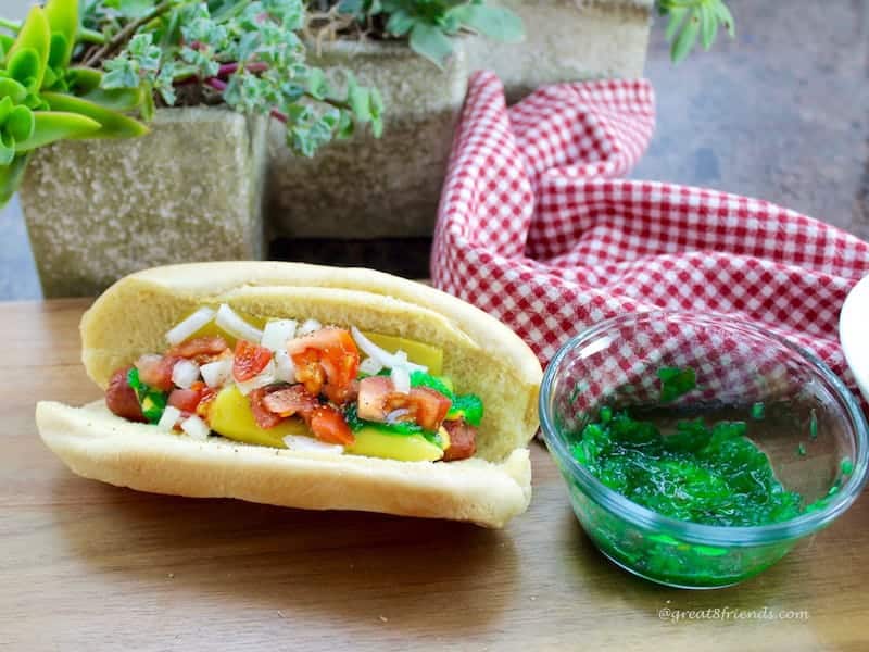 A fully loaded hot dog, Chicago style next to a bowl of bright green relish.