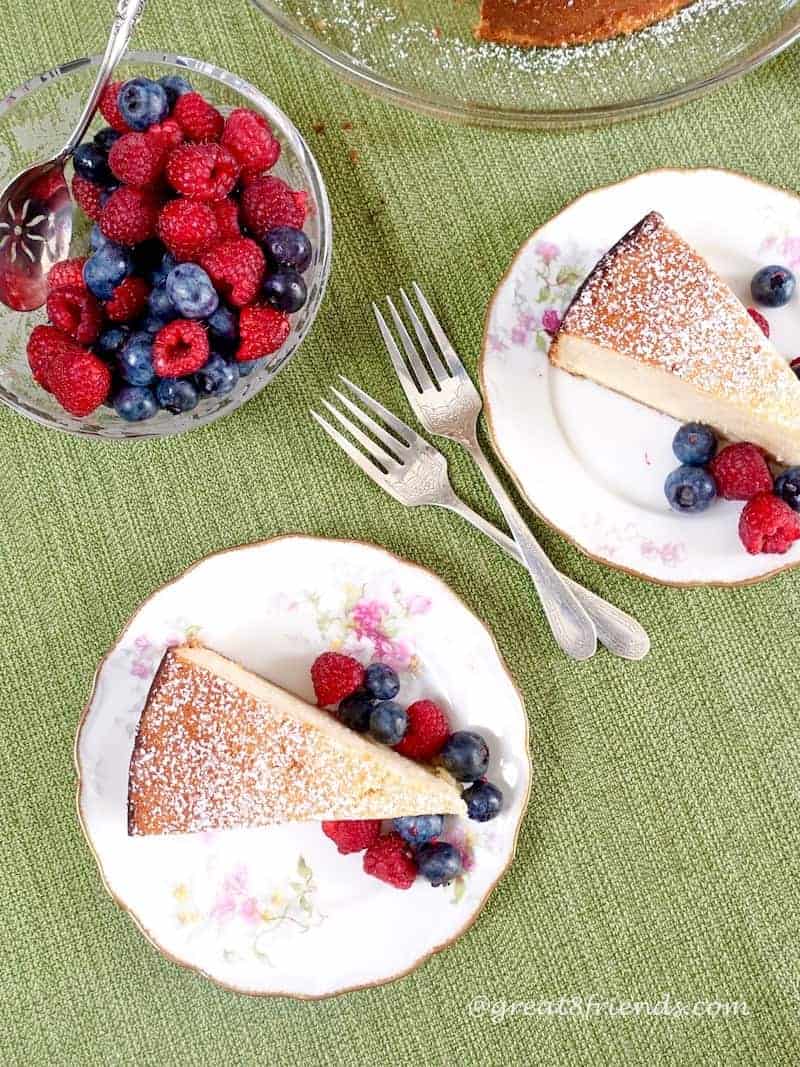 Overhead shot of 2 plates each with a slice of ricotta cake with berries. There are 2 forks in the center and a bowl of berries off to the side.