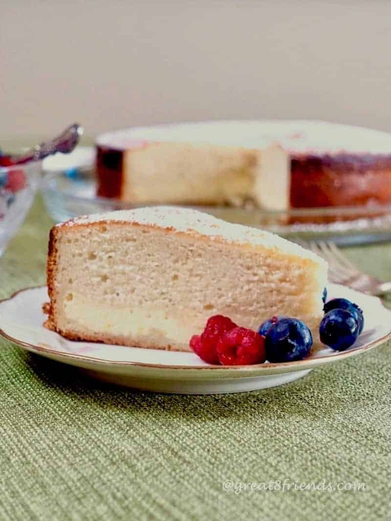 A slice of cake on a plate with berries. The larger cake is blurry in the background.