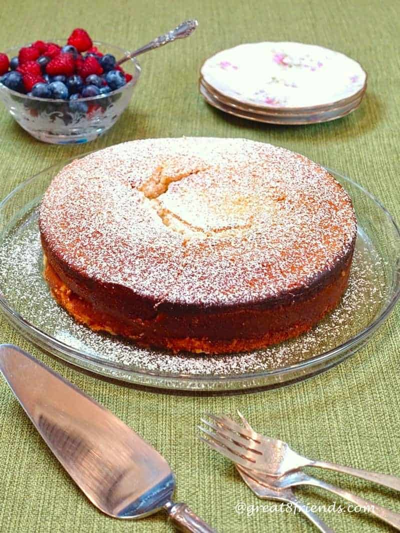 The whole ricotta Cake sprinkled with powdered sugar, with a small bowl of berries and a stack of 3 plates in the background.