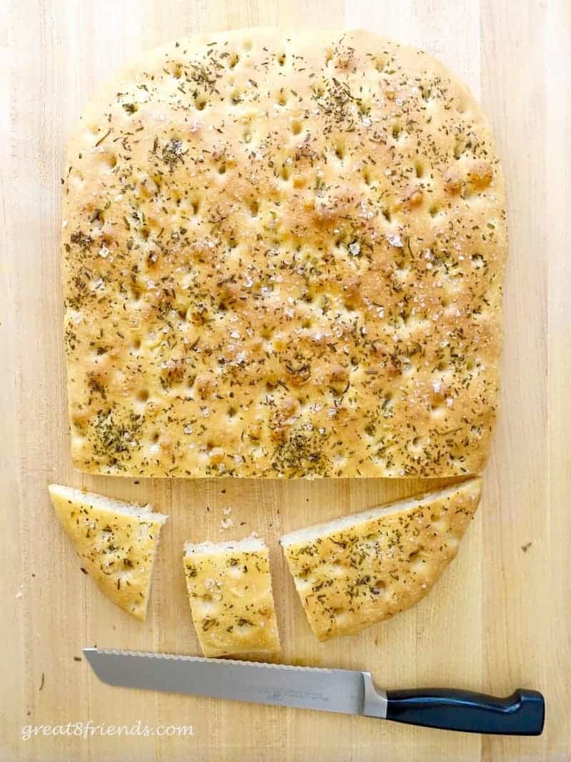 Rosemary Sea Salt Focaccia on wood cutting board with a serrated knife that has cut 3 pieces of the loaf.