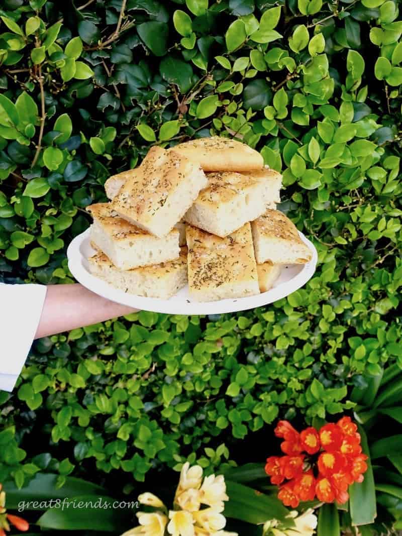 An arm holding a plate of Rosemary Sea Salt Focaccia against a background of bushes.